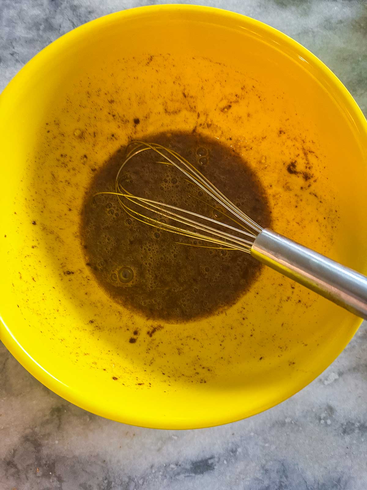 A metal whisk rests in a yellow bowl containing a brown, mixed liquid on a gray marble surface.