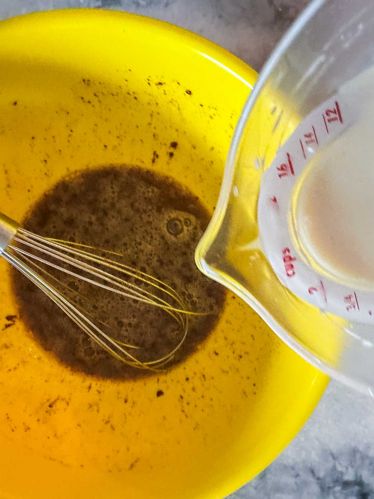 A whisk sits in a yellow bowl with a brown mixture while milk is being poured in from a measuring cup.