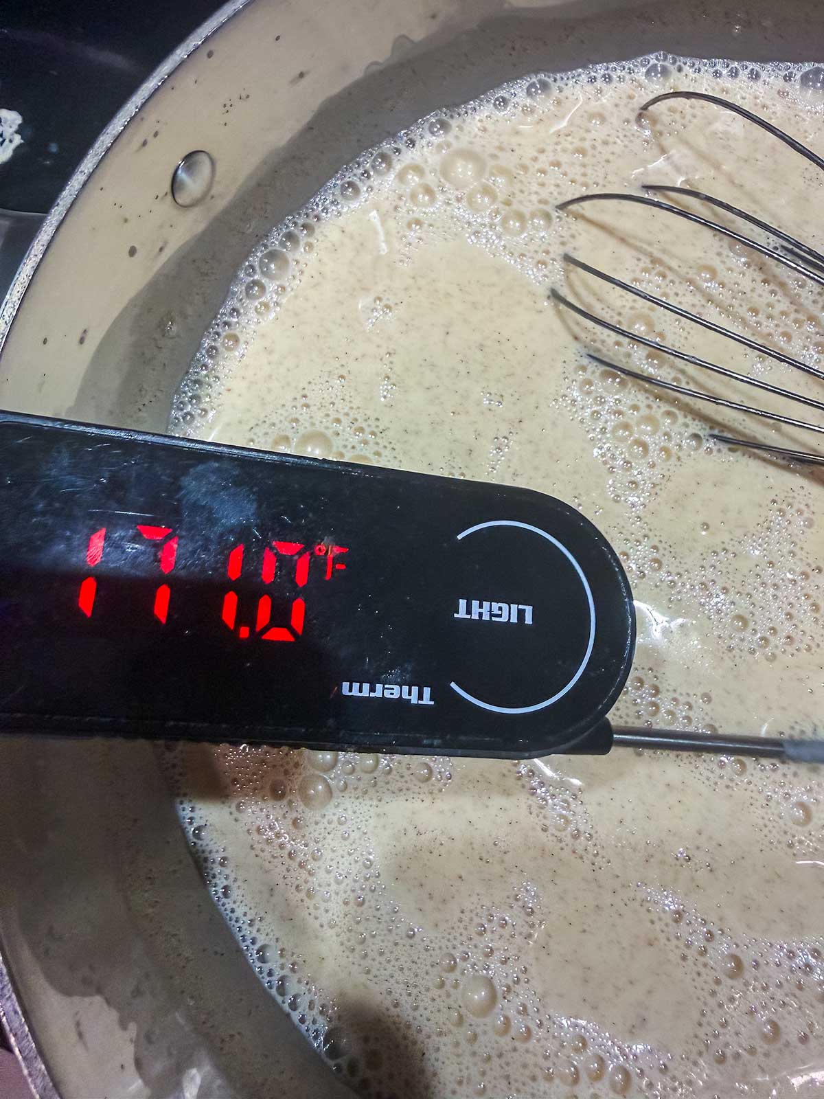 A digital thermometer displays 171.0°F while inserted in a pot of light brown, bubbly liquid. A metal whisk rests inside the pot, partially submerged in the mixture.