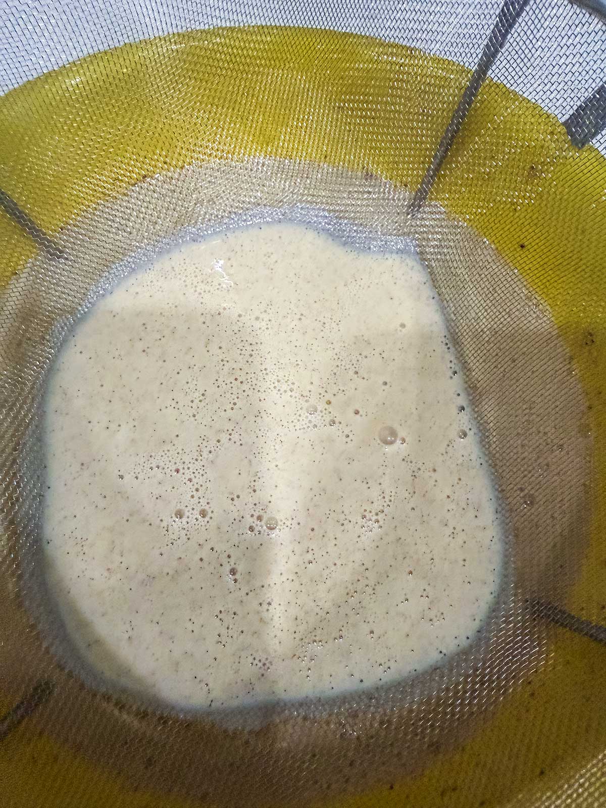 A close-up of a metal strainer containing a creamy beige liquid with bubbles, positioned over a yellow bowl.
