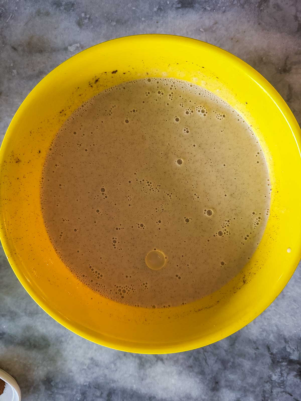 A yellow mixing bowl filled with a light brown batter sits on a gray marble surface. Small bubbles are visible on the surface of the batter.