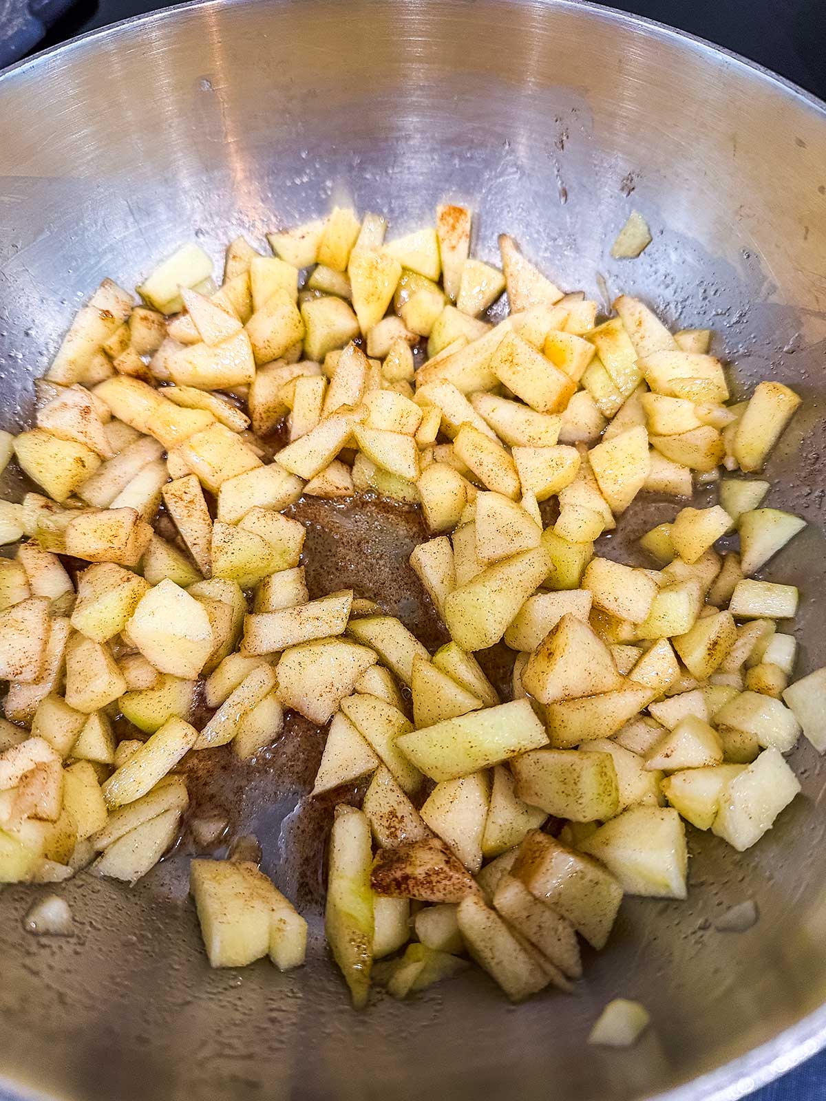 Chopped apples cooking in a metal pan, sprinkled with cinnamon and other spices, beginning to soften for a recipe.