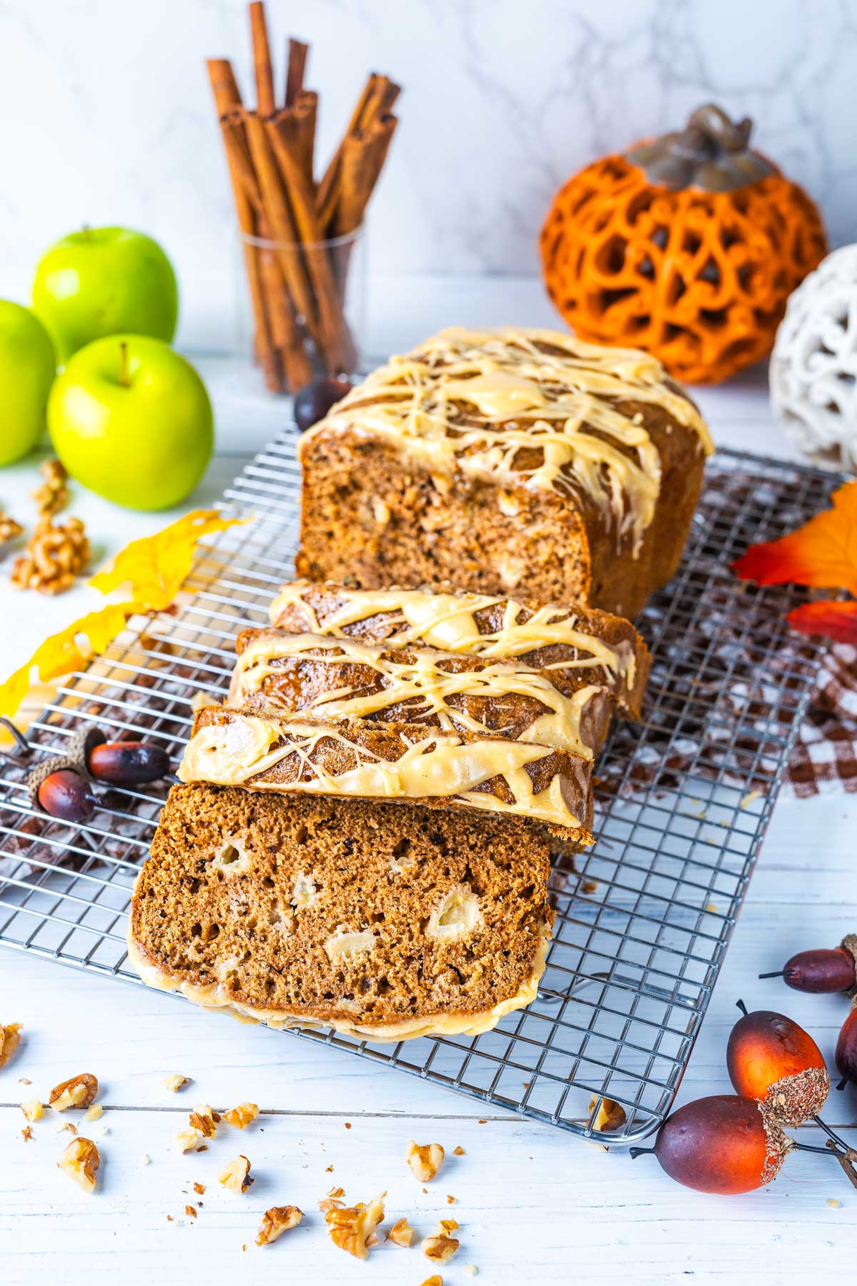A loaf of glazed apple bread with walnuts is sliced on a cooling rack. Green apples, cinnamon sticks, nuts, and autumn decorations are arranged around it on a white surface.