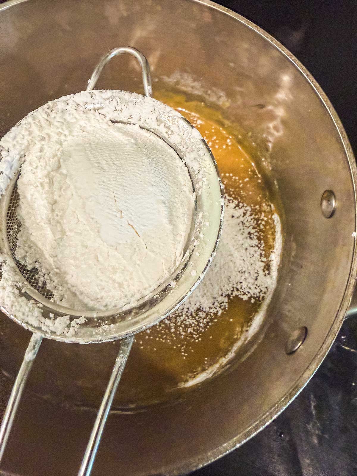 A metal sifter with flour is held over a mixing bowl containing a yellow batter, as flour is being sifted into the bowl.