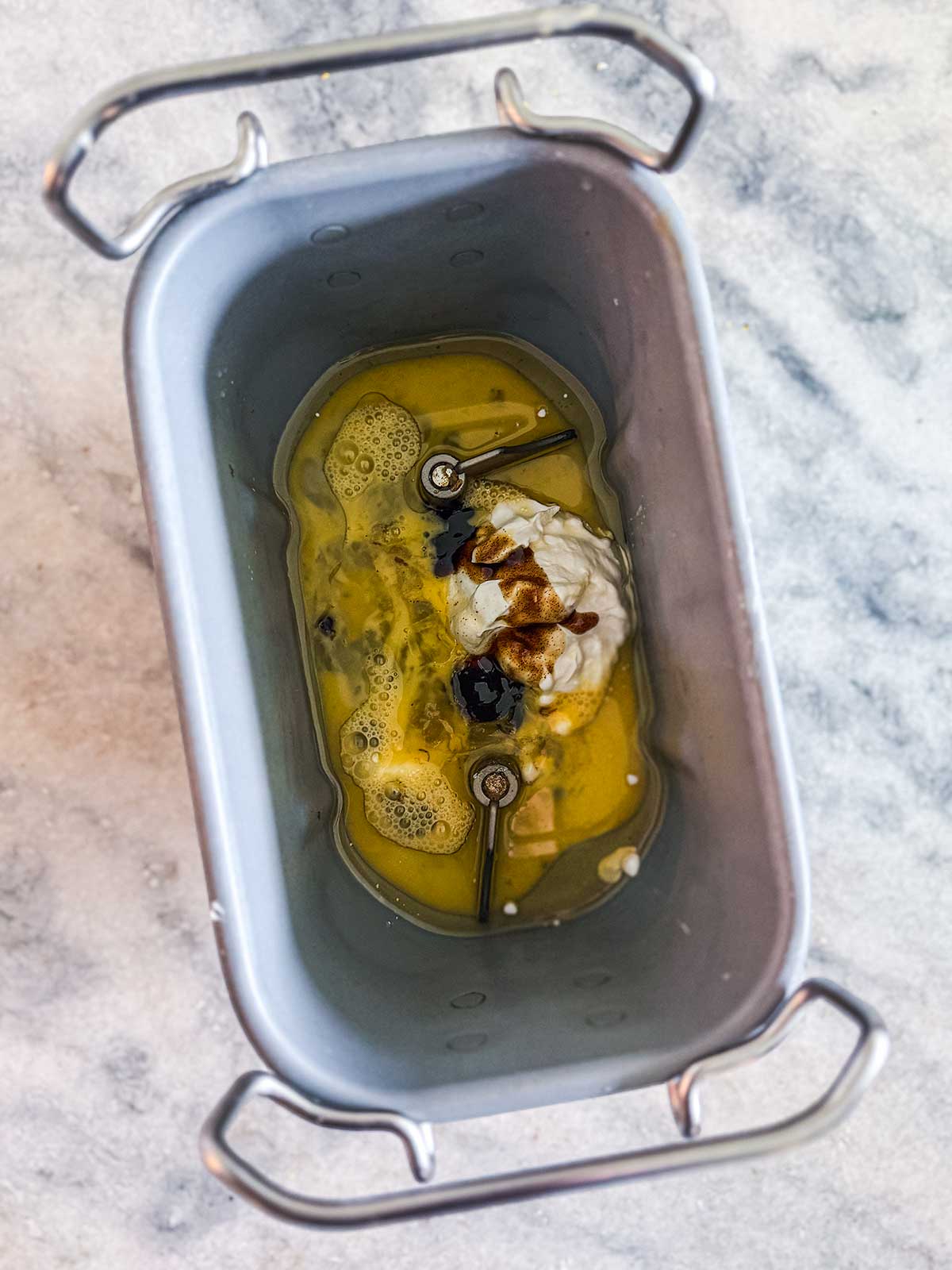 A bread machine pan with eggs, oil, flour, and dark liquid ingredients inside, viewed from above on a marble countertop.
