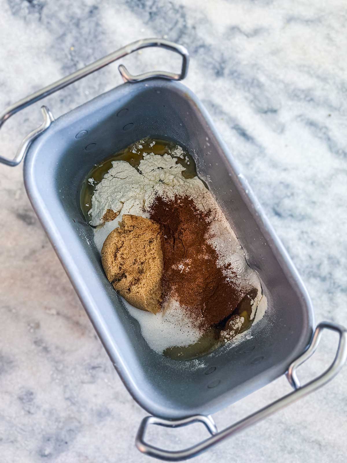 A bread machine pan filled with flour, brown sugar, cinnamon, white sugar, and eggs, ready for mixing, sits on a marble countertop.