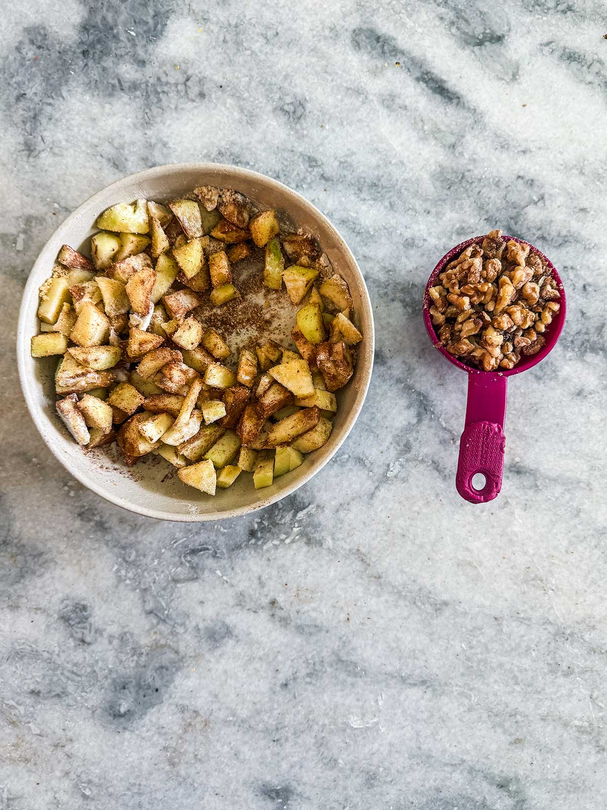 A bowl of chopped apples mixed with spices sits on a marble surface next to a pink measuring cup filled with chopped walnuts.