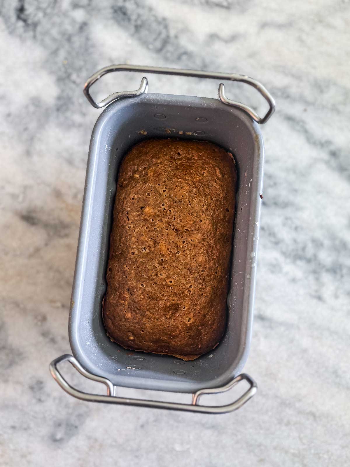 A loaf of baked bread sits in a metal loaf pan on a light, marbled countertop. The bread is brown and has a slightly bumpy surface.