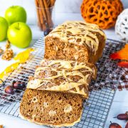 A loaf of spiced bread with icing sits on a cooling rack, partially sliced. Green apples, nuts, cinnamon sticks, acorns, and autumn leaves decorate the white surface around it.