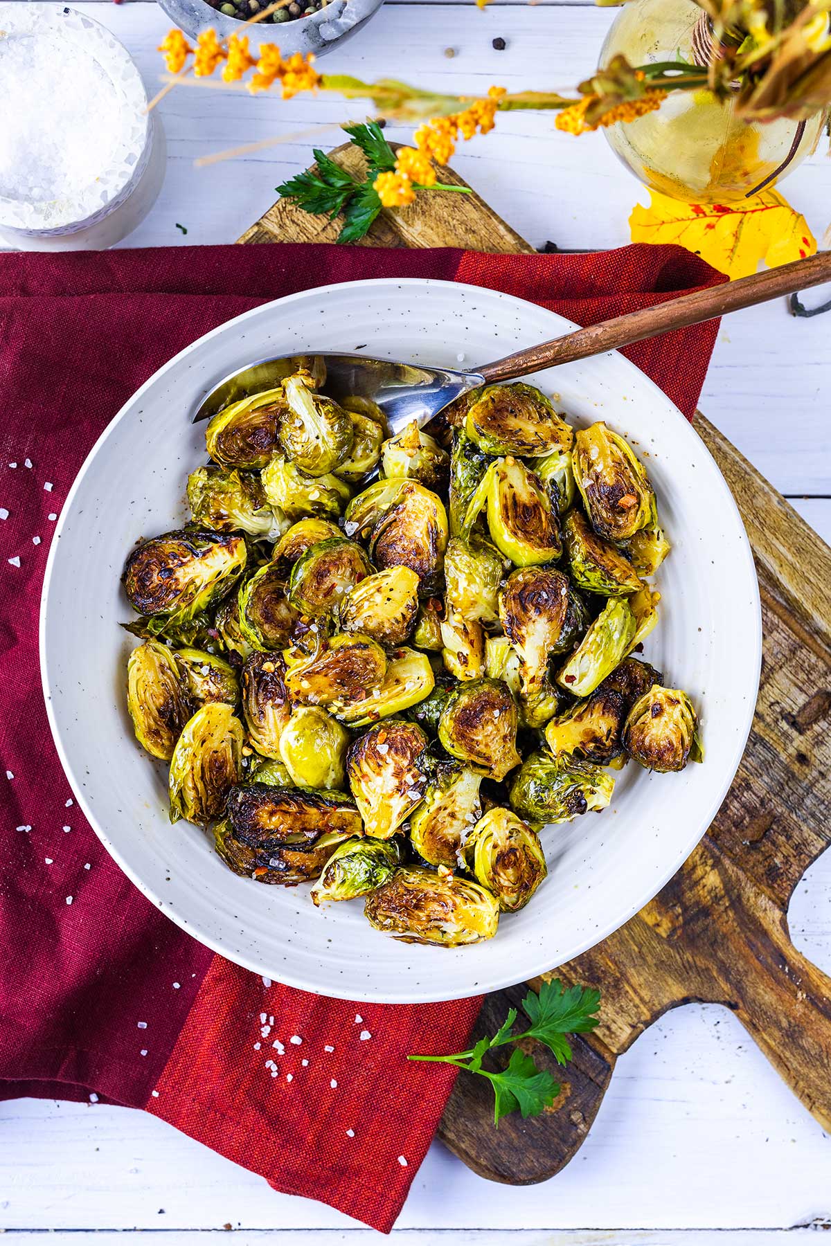 A white bowl filled with roasted Brussels sprouts sits on a red napkin and wooden board, sprinkled with coarse salt, with a fork in the bowl and some herbs nearby.