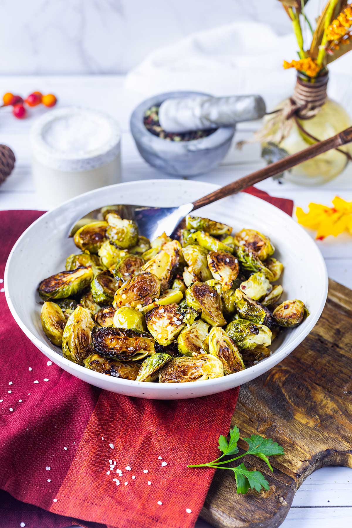 A bowl of roasted Brussels sprouts sits on a red napkin, topped with coarse salt. A spoon rests in the bowl. In the background are a vase, herbs, and a salt container.