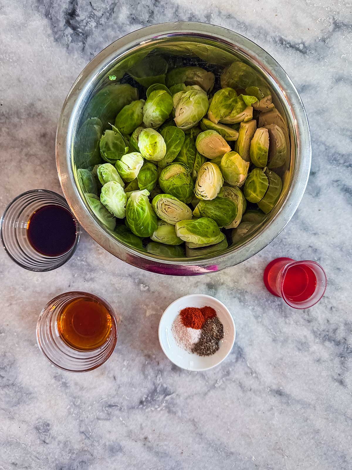 A metal bowl of halved Brussels sprouts, three small glasses with various liquids, and a small dish of mixed spices on a marble surface.