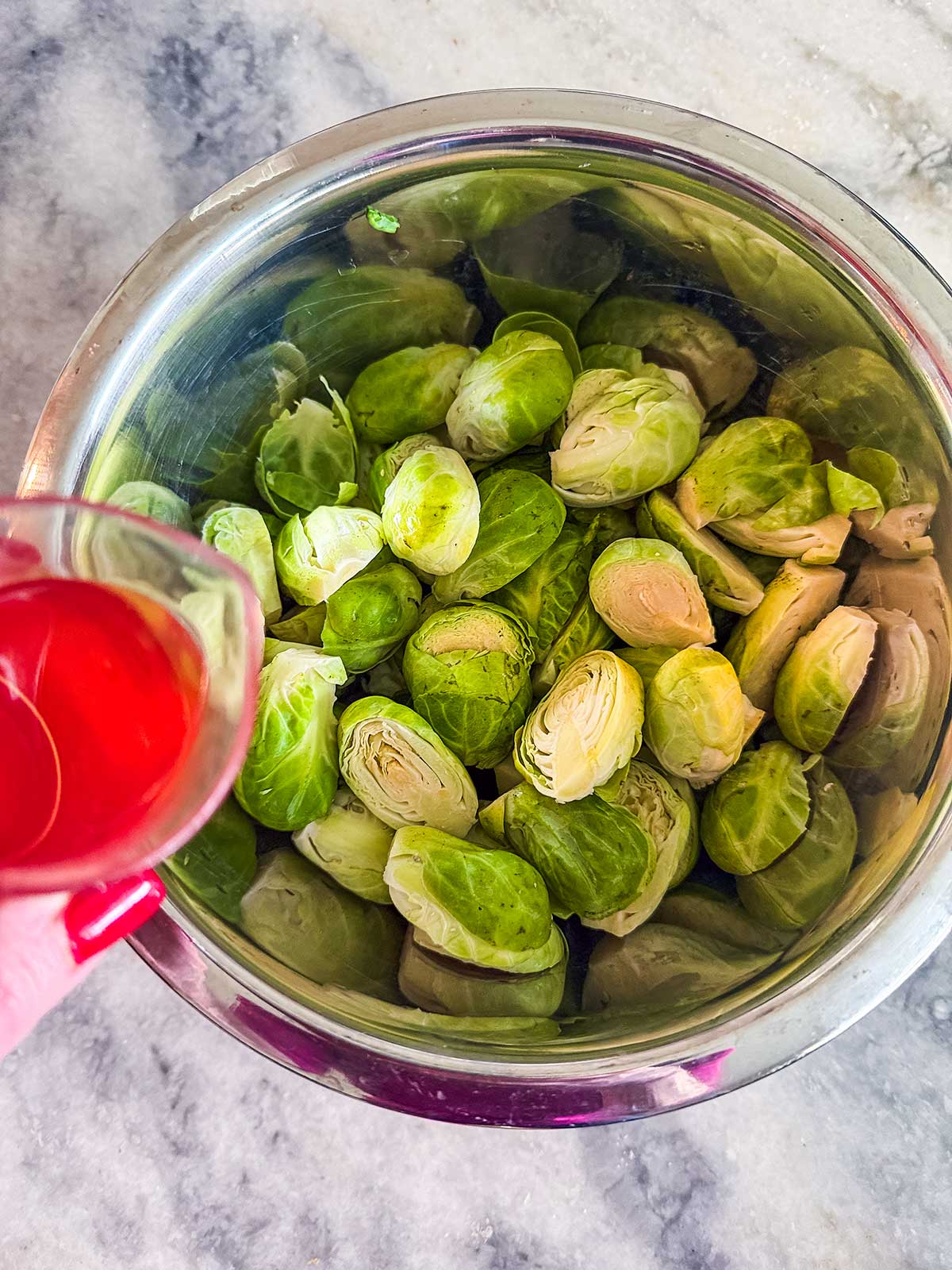 A hand holds a small glass of red liquid above a metal bowl filled with halved and whole Brussels sprouts on a marble countertop.