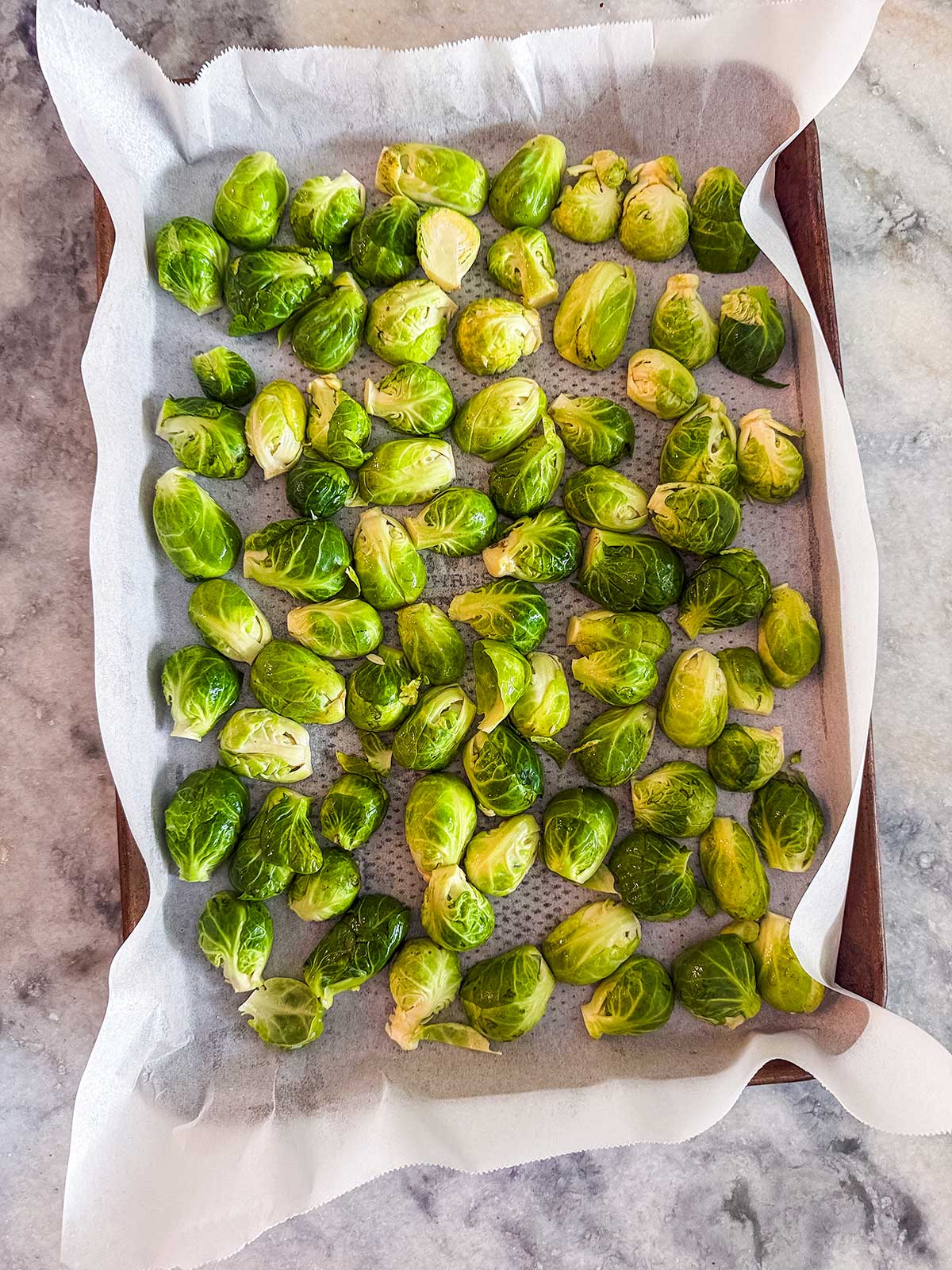 Brussels sprouts halves arranged on a parchment-lined baking tray, ready for roasting, on a marble countertop.