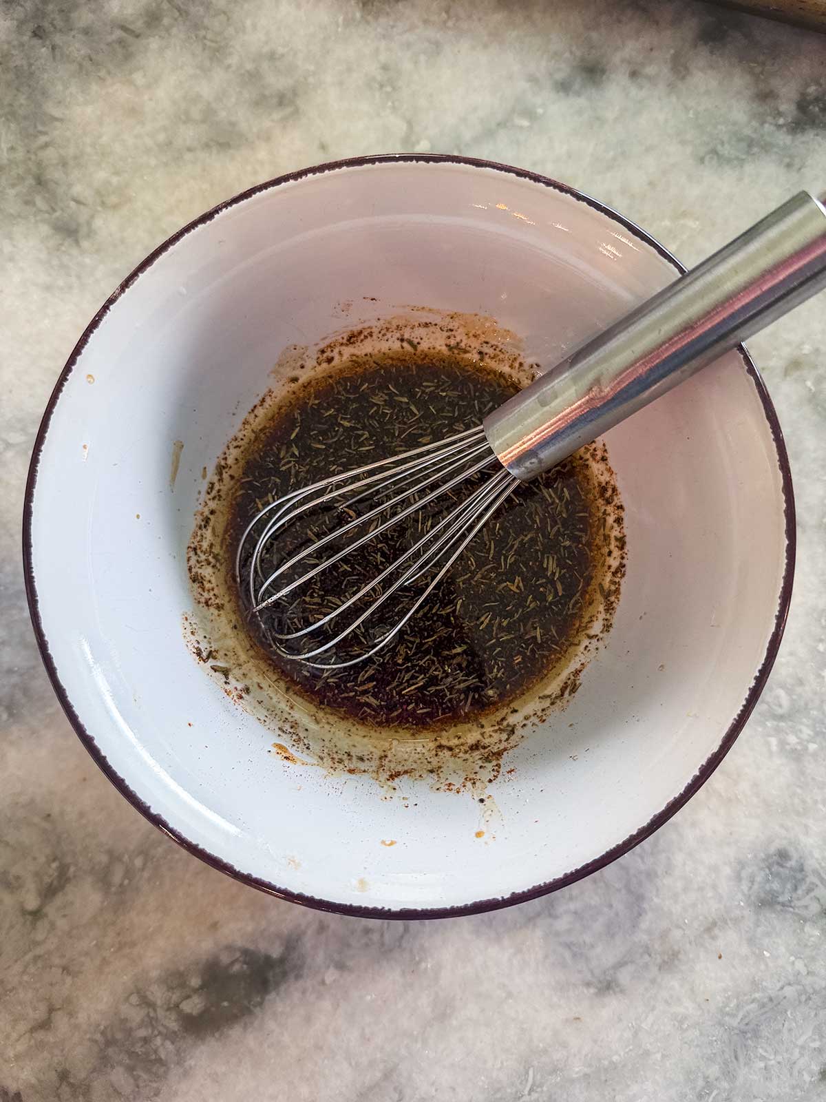 A metal whisk sits in a white bowl containing a dark, herb-filled liquid mixture on a marble countertop.