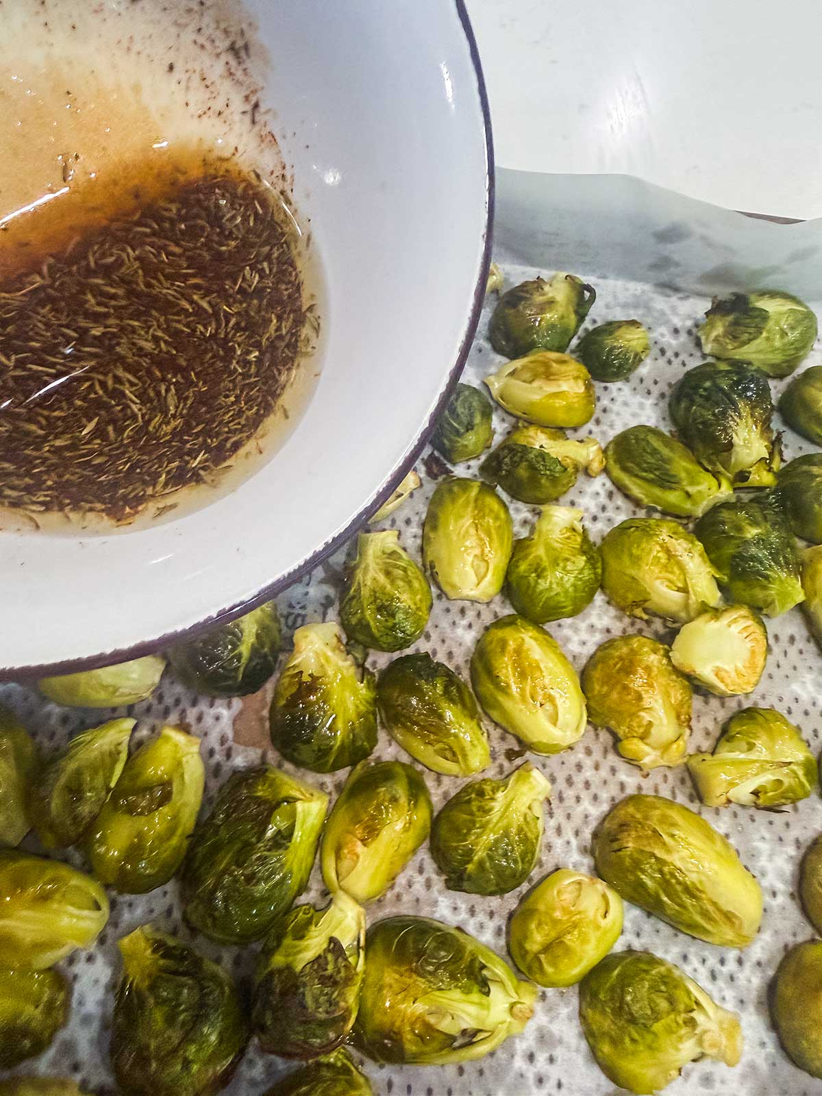 A baking sheet with roasted Brussels sprouts next to a bowl containing oil and dried herbs, likely for seasoning or tossing the vegetables.