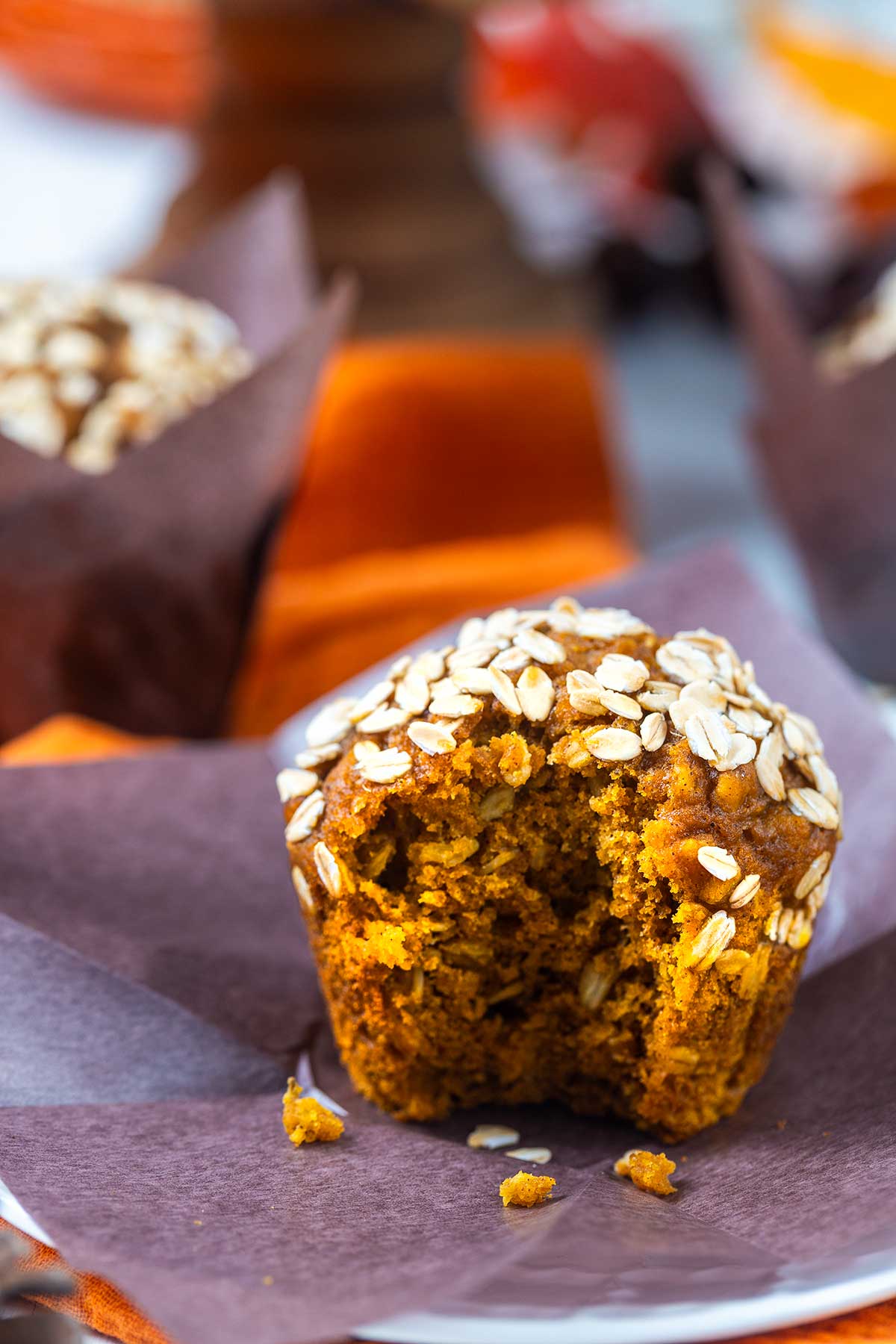A pumpkin muffin topped with oats sits on purple parchment paper, with a large bite taken out. Other muffins and orange fabric are blurred in the background.