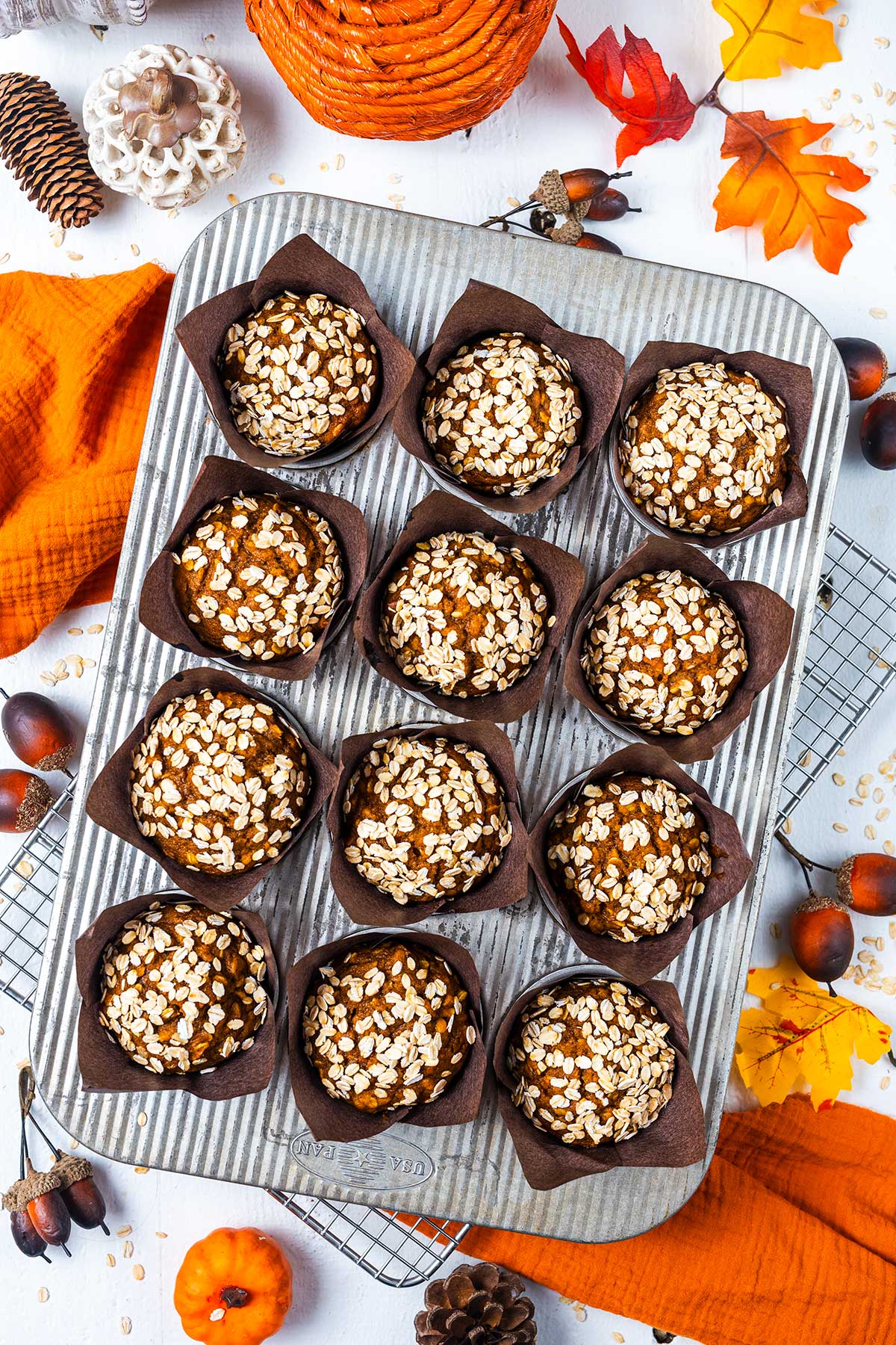 A muffin tin holds twelve oat-topped muffins in brown wrappers. Autumn leaves, pinecones, acorns, orange cloth, and mini pumpkins surround the tin, creating a festive fall atmosphere.