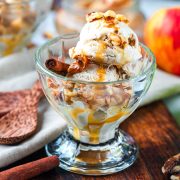 A glass dish with scoops of ice cream topped with chopped nuts and caramel sauce, garnished with a cinnamon stick. An apple and wooden spoon are visible in the background.