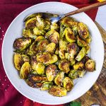 A white bowl filled with roasted Brussels sprouts, some pieces browned and crispy, sits on a red cloth with a spoon resting in the bowl.