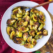 A white bowl filled with roasted Brussels sprouts, some pieces browned and crispy, sits on a red cloth with a spoon resting in the bowl.