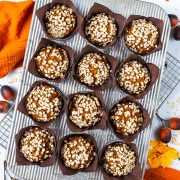 Twelve pumpkin muffins topped with oats in brown paper liners sit on a metal tray. Autumn leaves, acorns, a wire rack, and an orange cloth are visible around the tray.