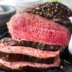 A close-up of a medium-rare roast beef, sliced to show its juicy pink center and seasoned crust, topped with coarse salt and herbs.
