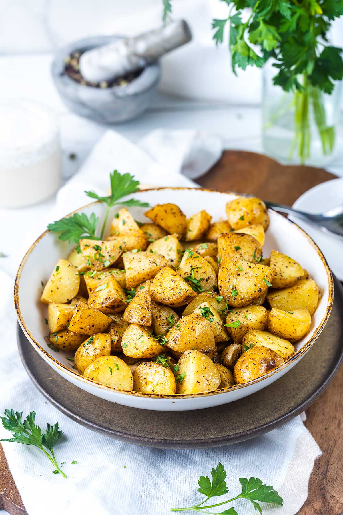 A bowl of roasted potato chunks garnished with chopped parsley sits on a plate, with fresh parsley and a garlic mortar visible in the background.