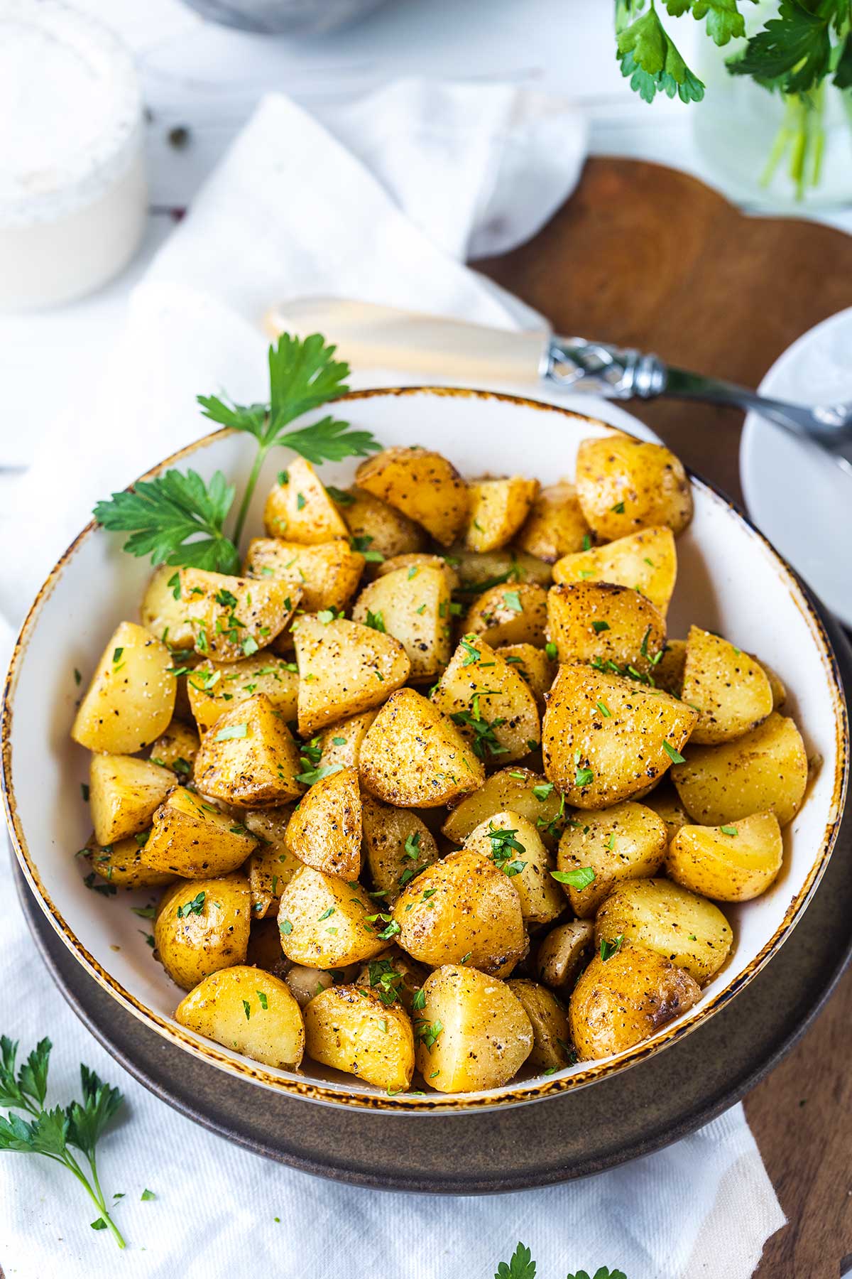 A bowl of roasted potato cubes garnished with chopped parsley sits on a white napkin, with a fork and sprigs of fresh parsley nearby.