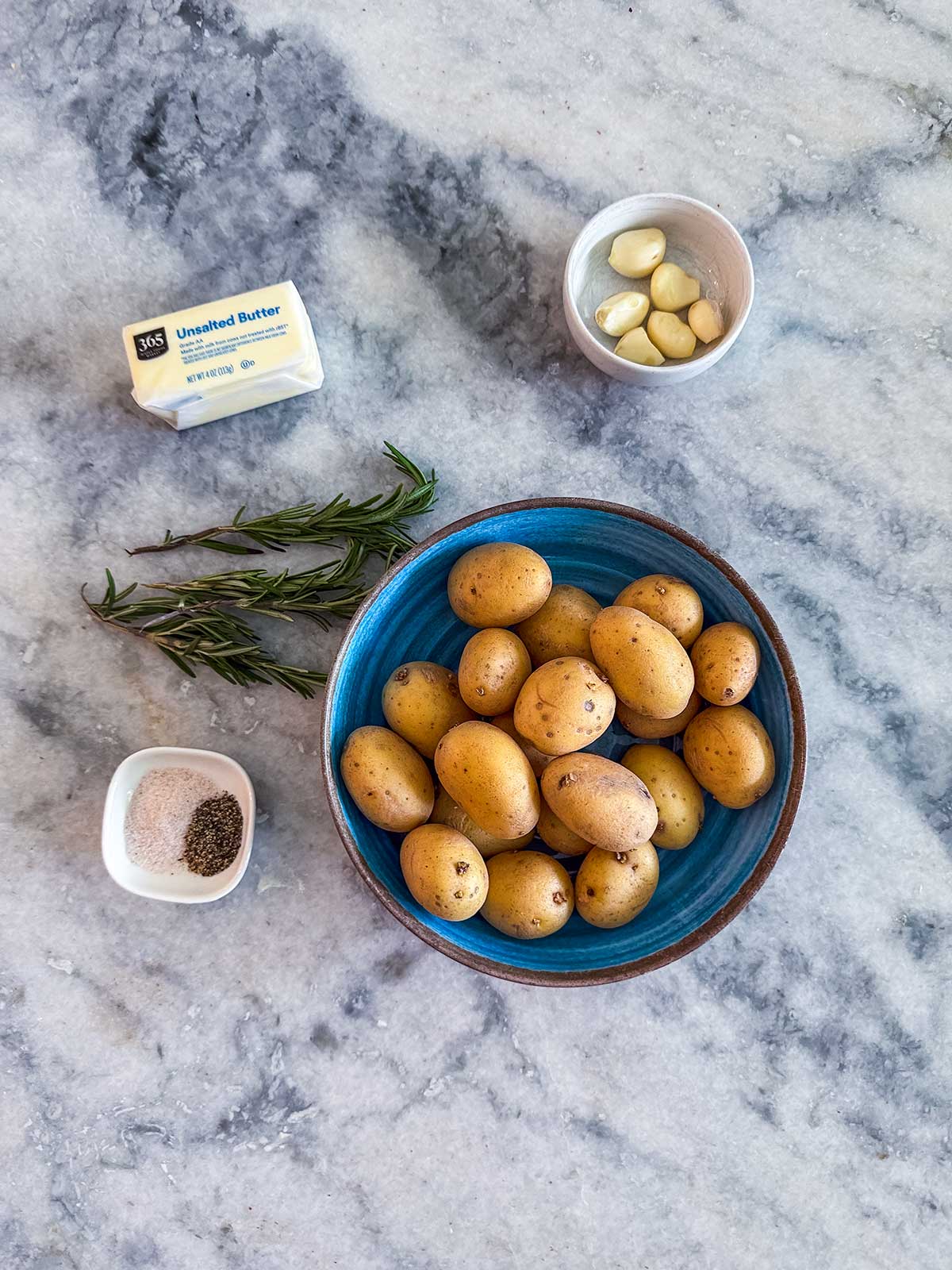 A blue bowl of baby potatoes sits on a marble counter. Surrounding it are sprigs of rosemary, a stick of unsalted butter, garlic cloves in a small bowl, and separate dishes of salt and pepper.