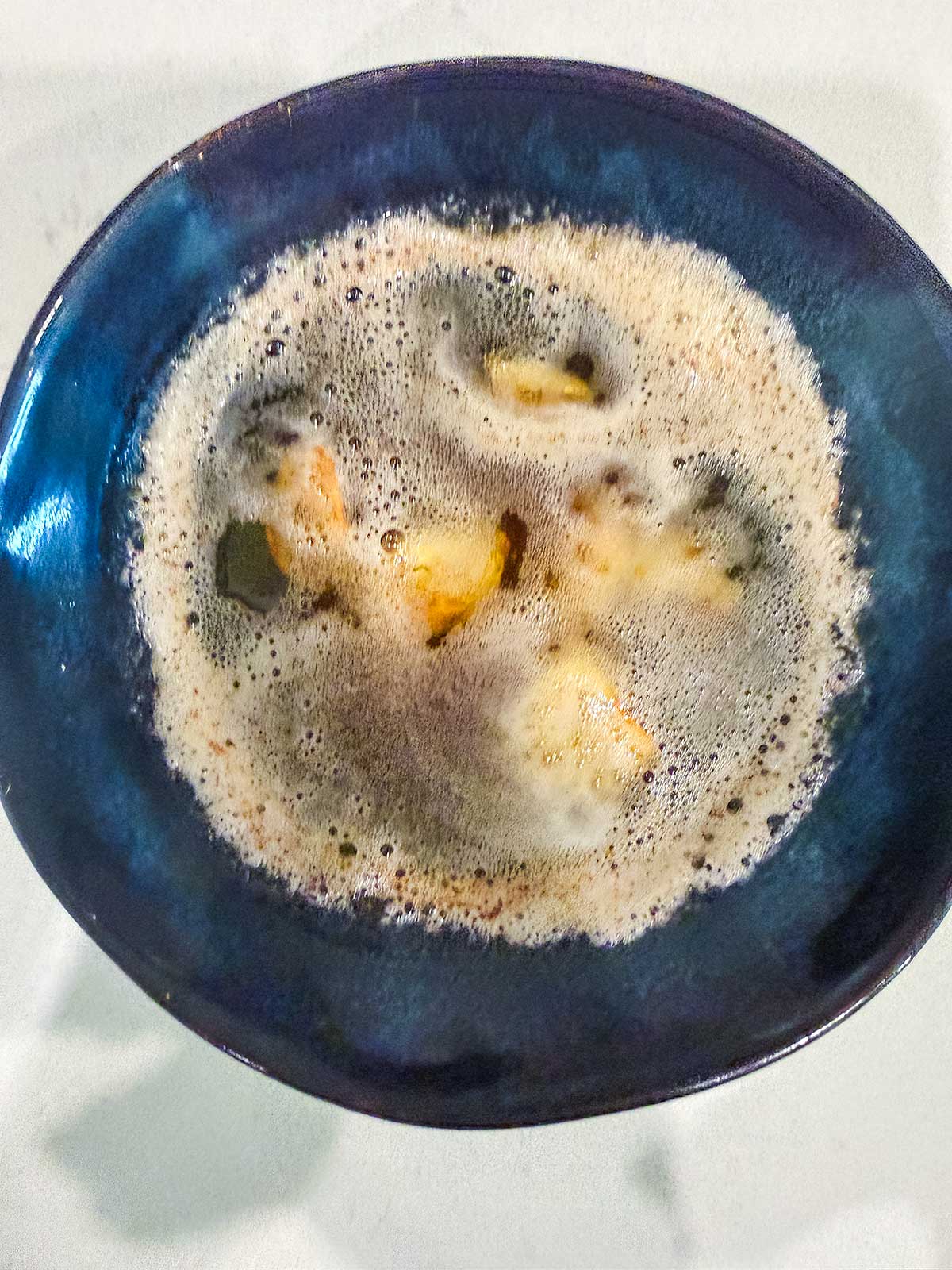 A blue bowl filled with a frothy, bubbly liquid and chunks of yellow fruit or vegetable, viewed from above on a light-colored surface.
