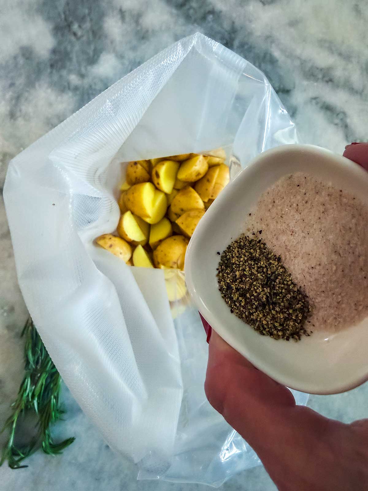 A hand holds a small bowl of salt and pepper over a vacuum-seal bag filled with cut potatoes. A sprig of rosemary and a marble countertop are visible in the background.