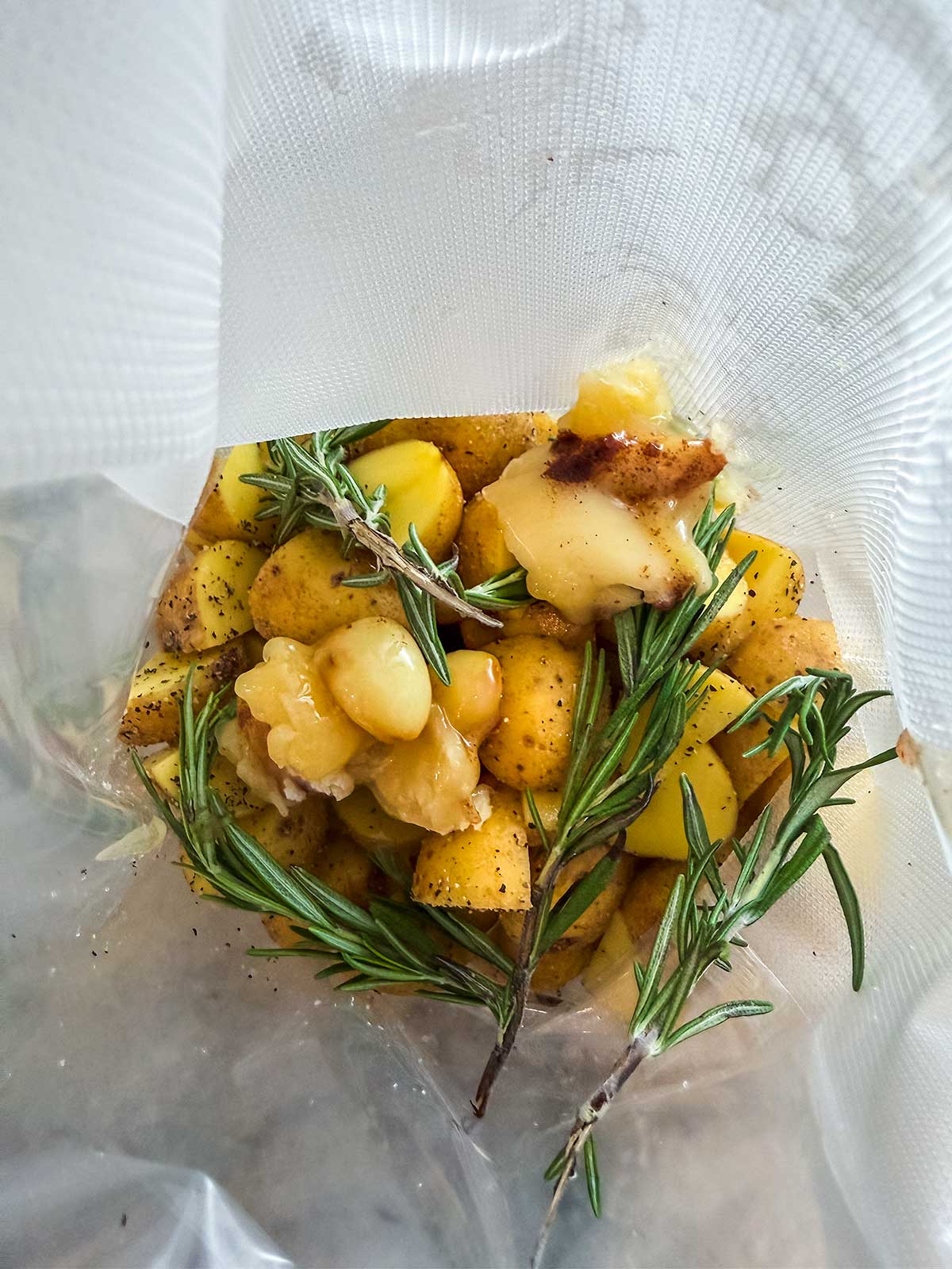 Small yellow potatoes, sprigs of fresh rosemary, and pieces of butter are inside a clear vacuum-sealed plastic bag, ready for sous vide cooking.