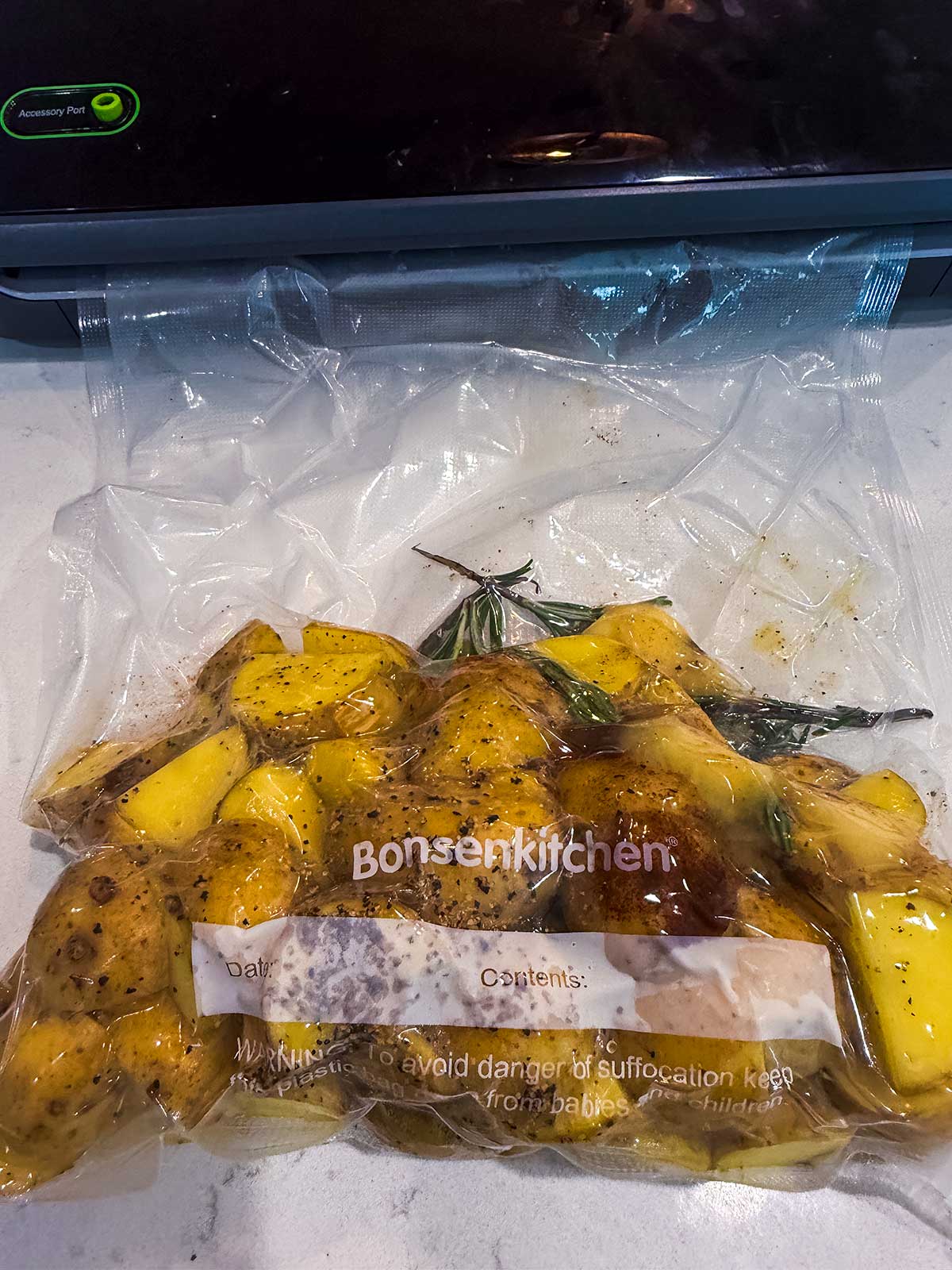 Vacuum-sealed bag with seasoned yellow potatoes and rosemary, labeled Bonsenkitchen, placed on a white surface near a vacuum sealer machine.