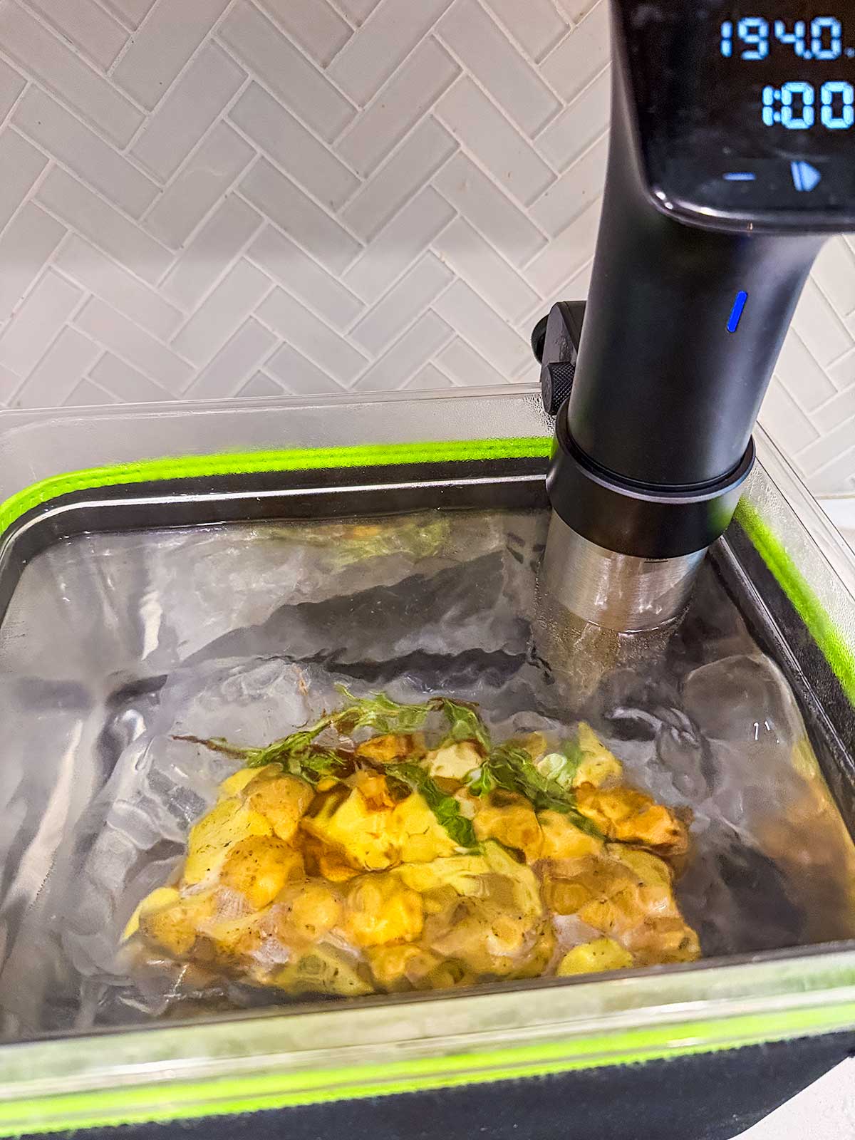 A sous vide machine circulates water in a clear container with sealed food, including herbs and seasonings, immersed inside. The temperature display reads 134.0°F. The background has white subway tile.