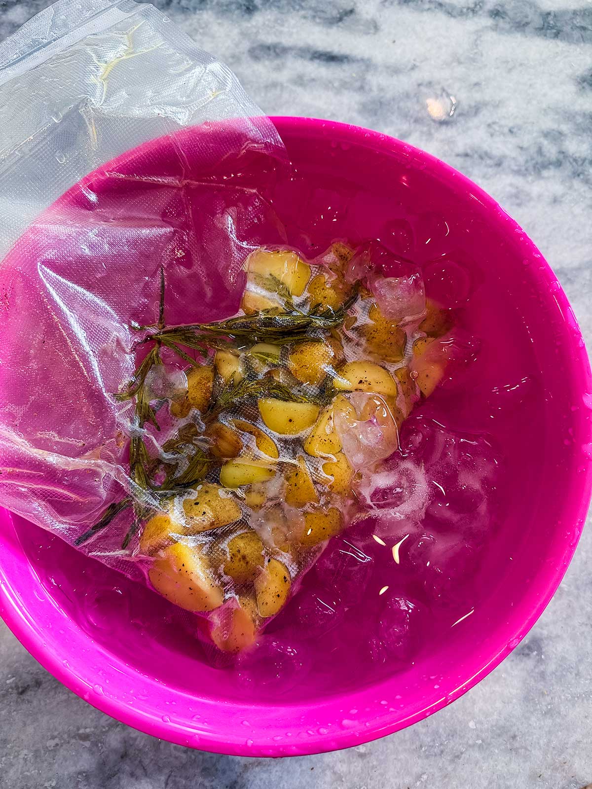 A vacuum-sealed bag with garlic, herbs, and oil is cooling in ice water inside a bright pink bowl on a marble countertop.