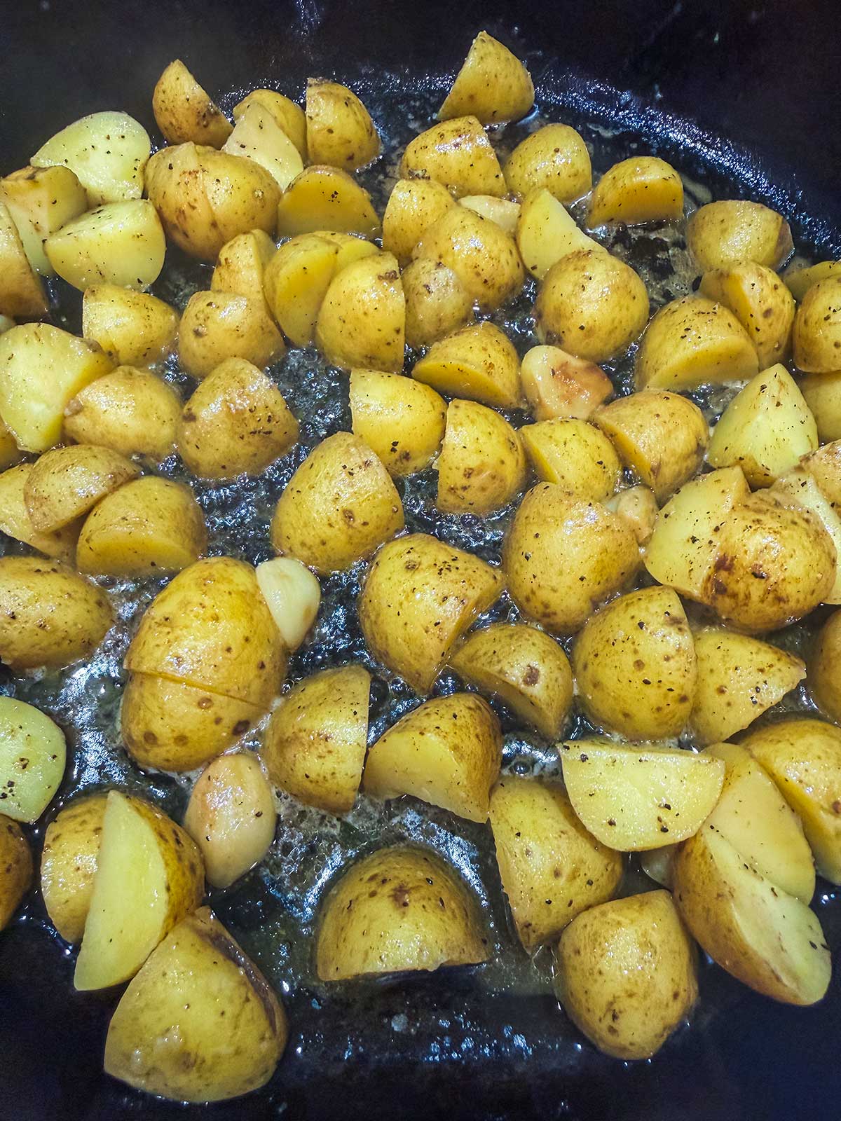 Quartered yellow potatoes cooking in a skillet with oil and seasonings, sizzling and browning on the surface.