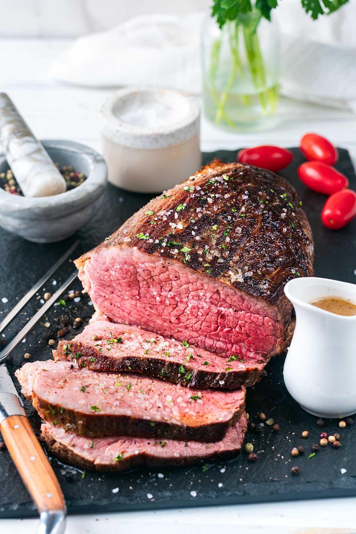 A sliced roast beef is displayed on a slate board with cherry tomatoes, a knife, peppercorns, a small gravy boat, and a mortar and pestle nearby. The beef is medium-rare and topped with herbs and salt.