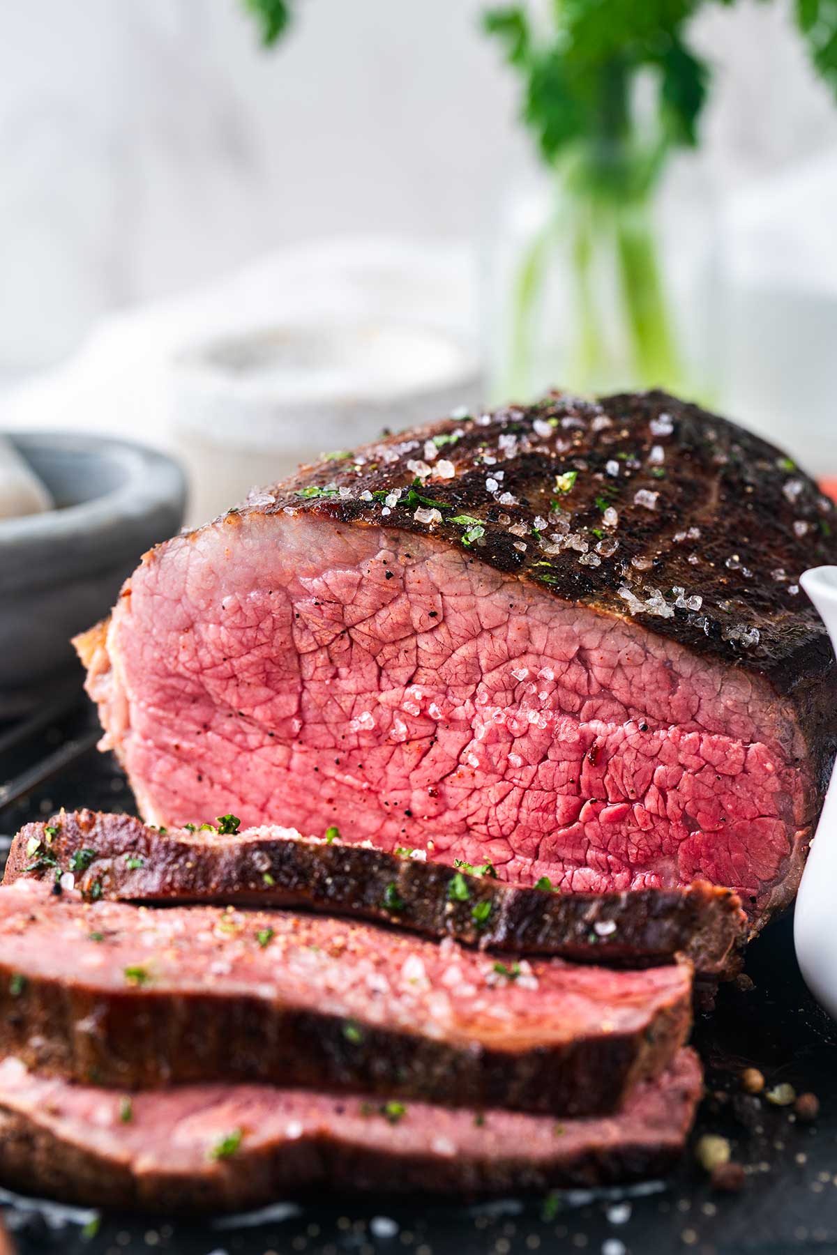 Close-up of a sliced, medium-rare roast beef with a browned crust, sprinkled with coarse salt and herbs. A blurred background shows a white bowl and a glass jar with green leaves.