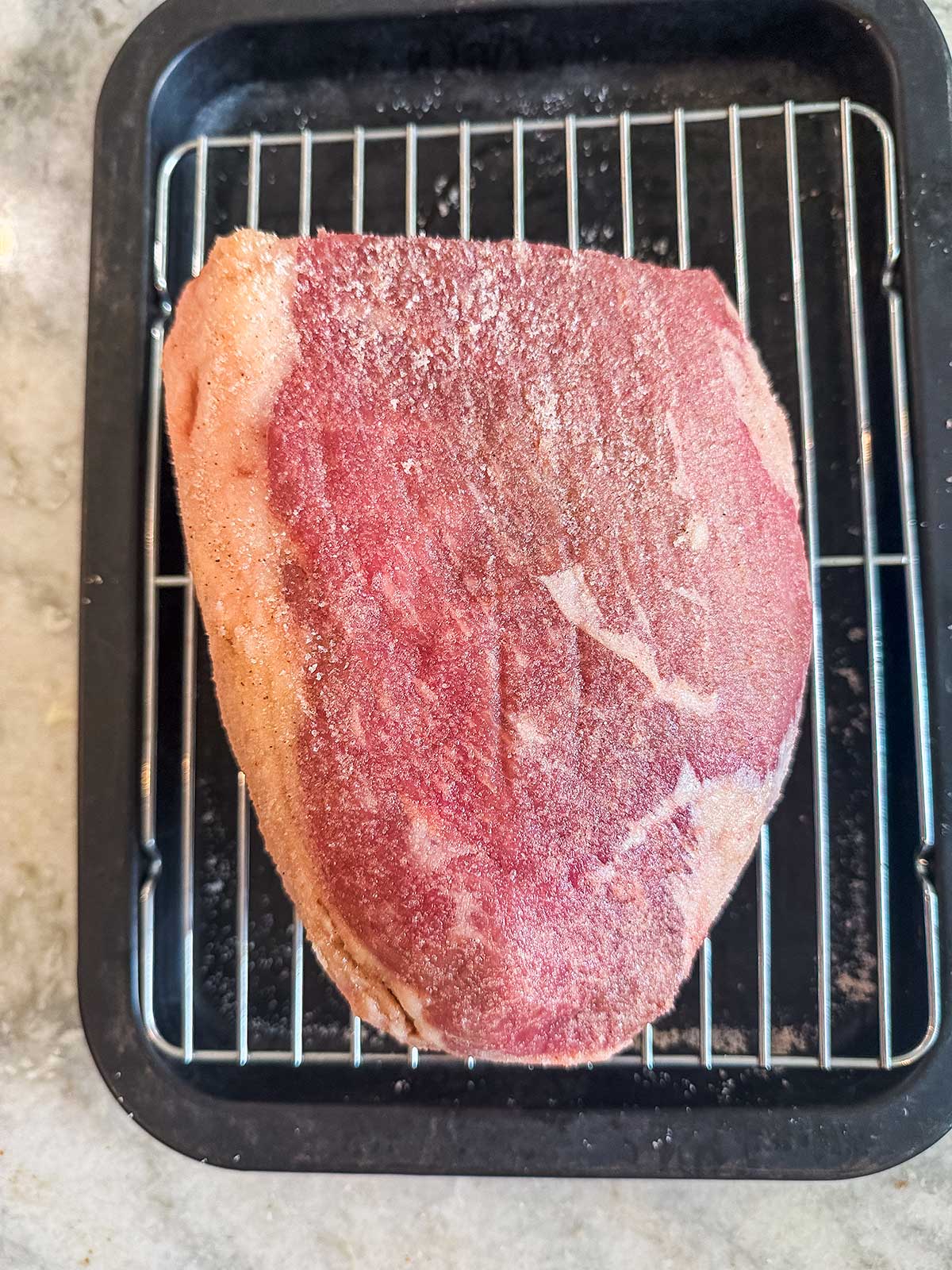 A large, raw roast beef covered with seasoning sits on a metal rack inside a black roasting pan, ready for cooking.