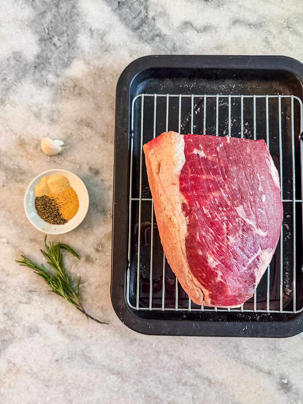 Raw beef on a wire rack in a black roasting pan, with a bowl of spices, a garlic clove, and a sprig of rosemary on a marble countertop.