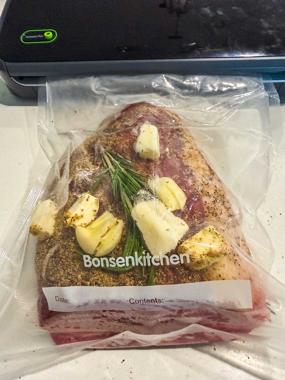 A vacuum-sealed bag with seasoned raw meat, fresh rosemary, garlic cloves, and butter pieces, labeled Bonsenkitchen, sits on a white surface with a food vacuum sealer machine in the background.
