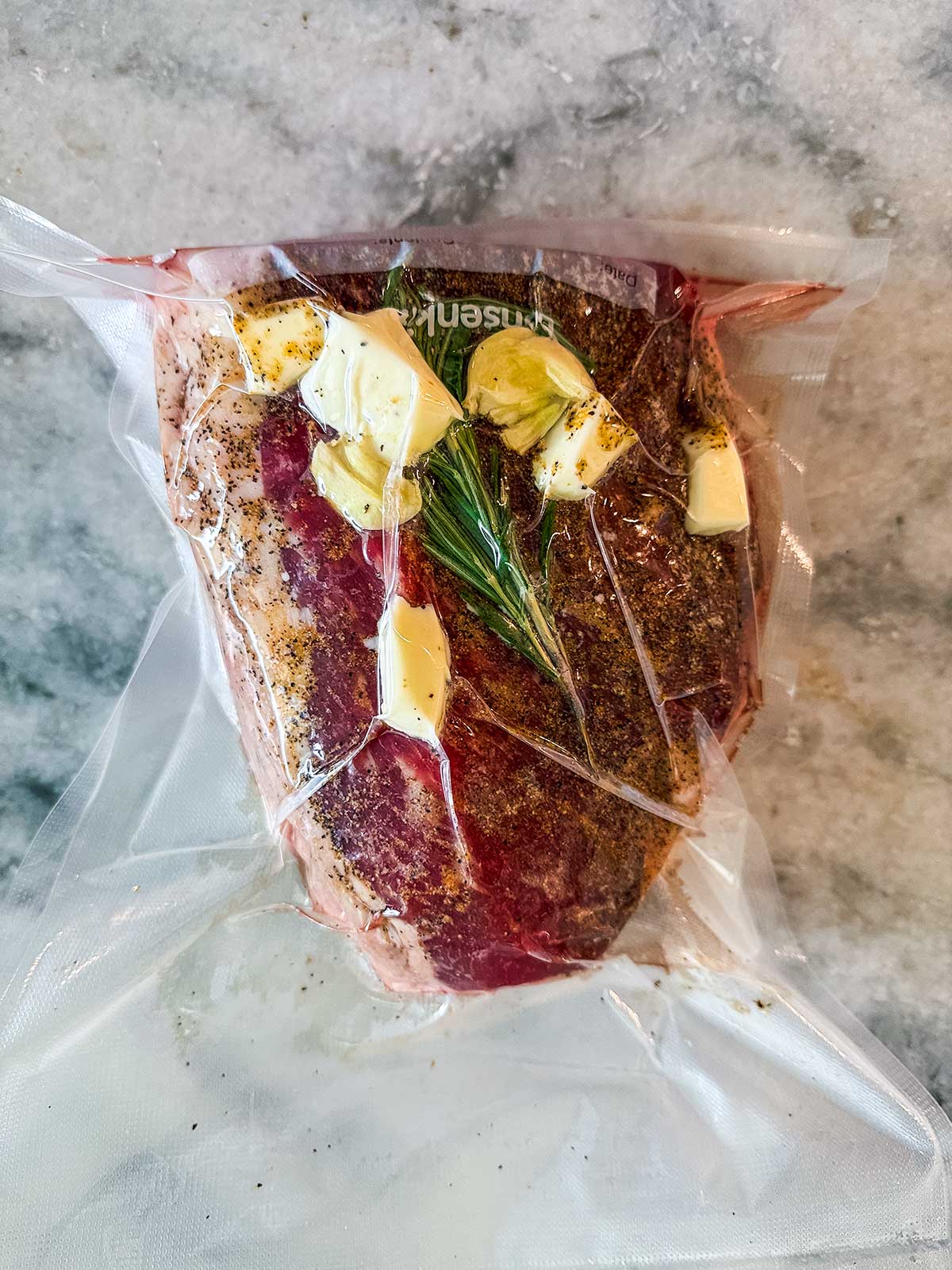 A vacuum-sealed raw steak is seasoned with pepper, topped with butter, garlic, and a sprig of rosemary, resting on a marbled countertop.