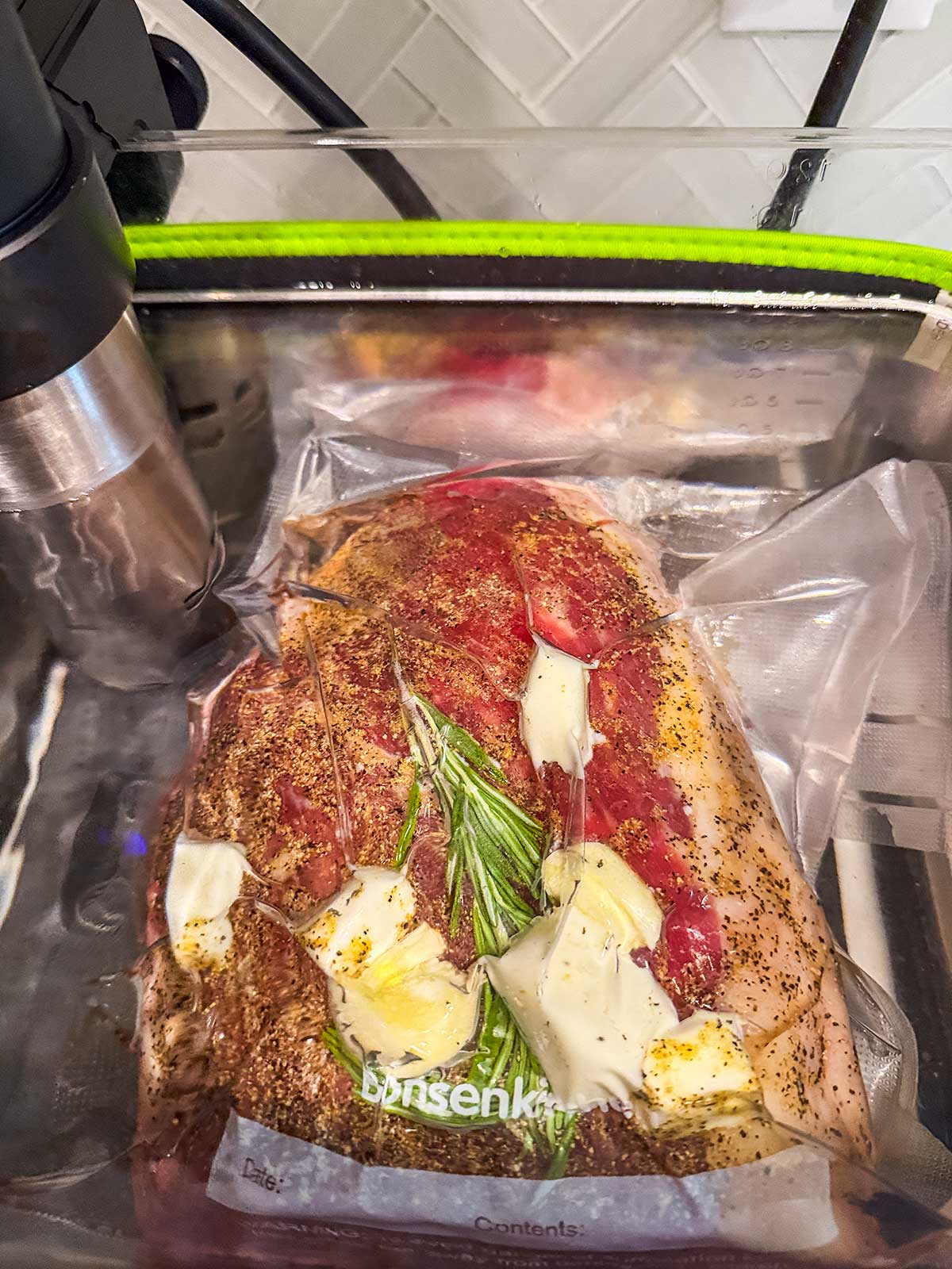 Vacuum-sealed steak with herbs, garlic, and butter cooking in a sous vide water bath, with a sous vide immersion circulator attached to the side of the clear container.