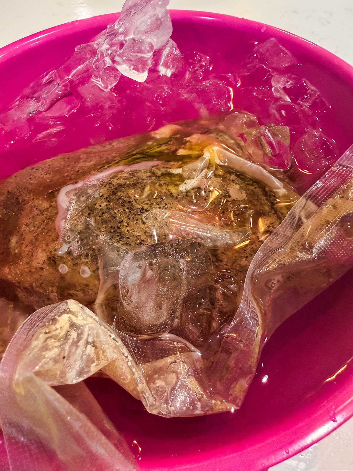 A vacuum-sealed bag with seasoned meat is submerged in a pink bowl filled with ice water, likely for cooling after sous vide cooking.
