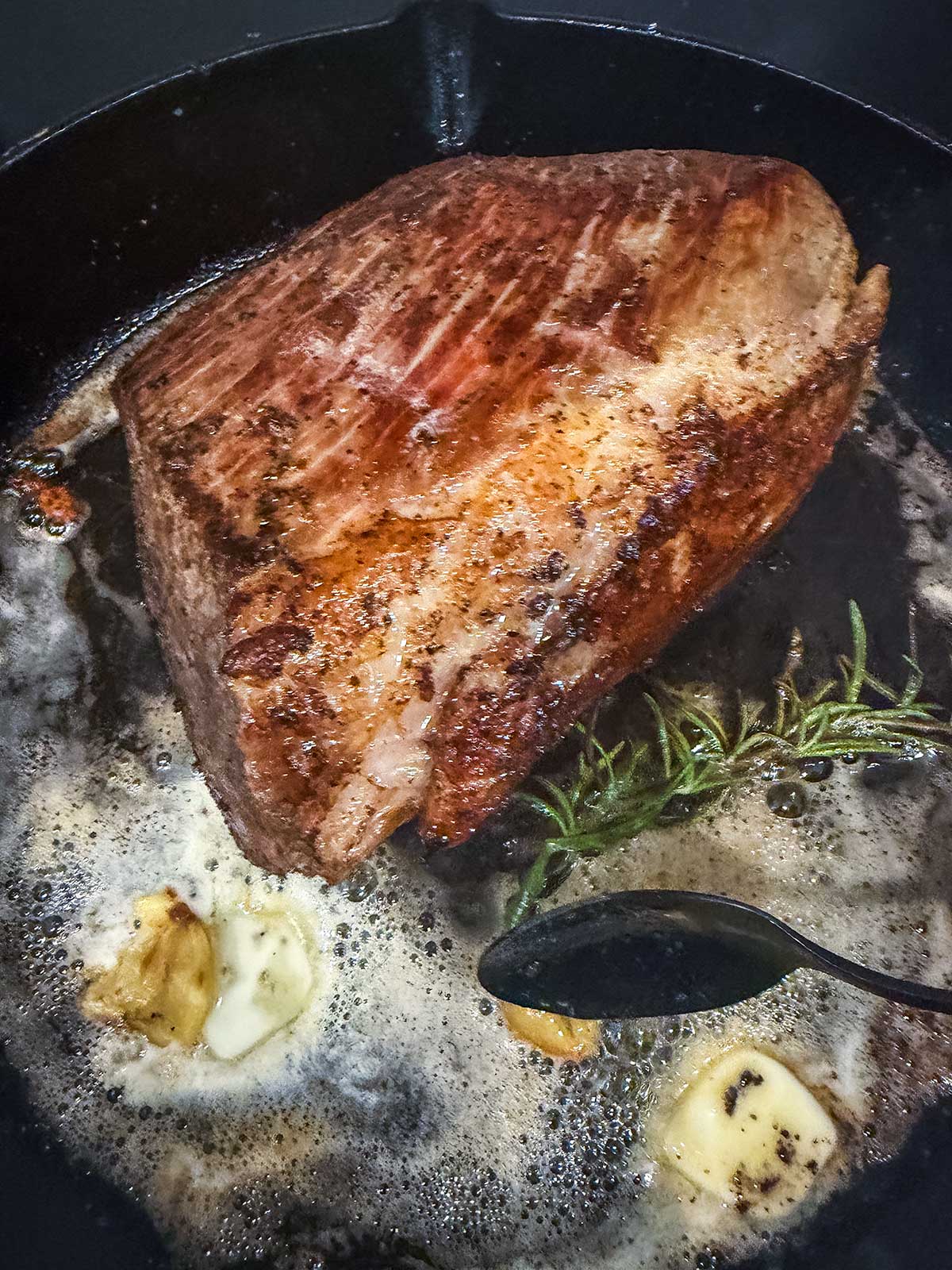 A seared steak cooks in a cast iron pan with melted butter, garlic cloves, and a sprig of rosemary. A spoon is shown basting the steak with the butter.