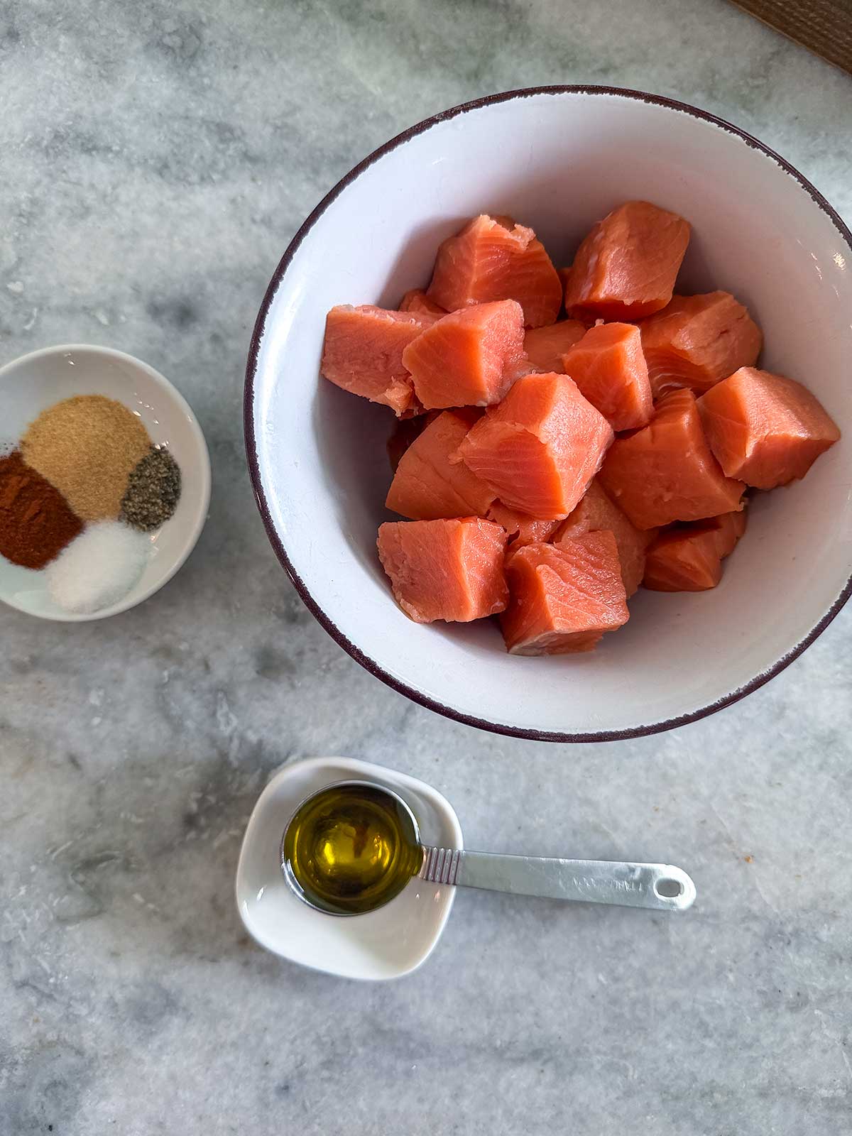 A bowl of cubed salmon—perfect for making Salmon Bites or Honey Garlic Salmon—is set beside a small dish of mixed spices and a small bowl with a spoonful of olive oil on a gray countertop, ready for the air fryer.