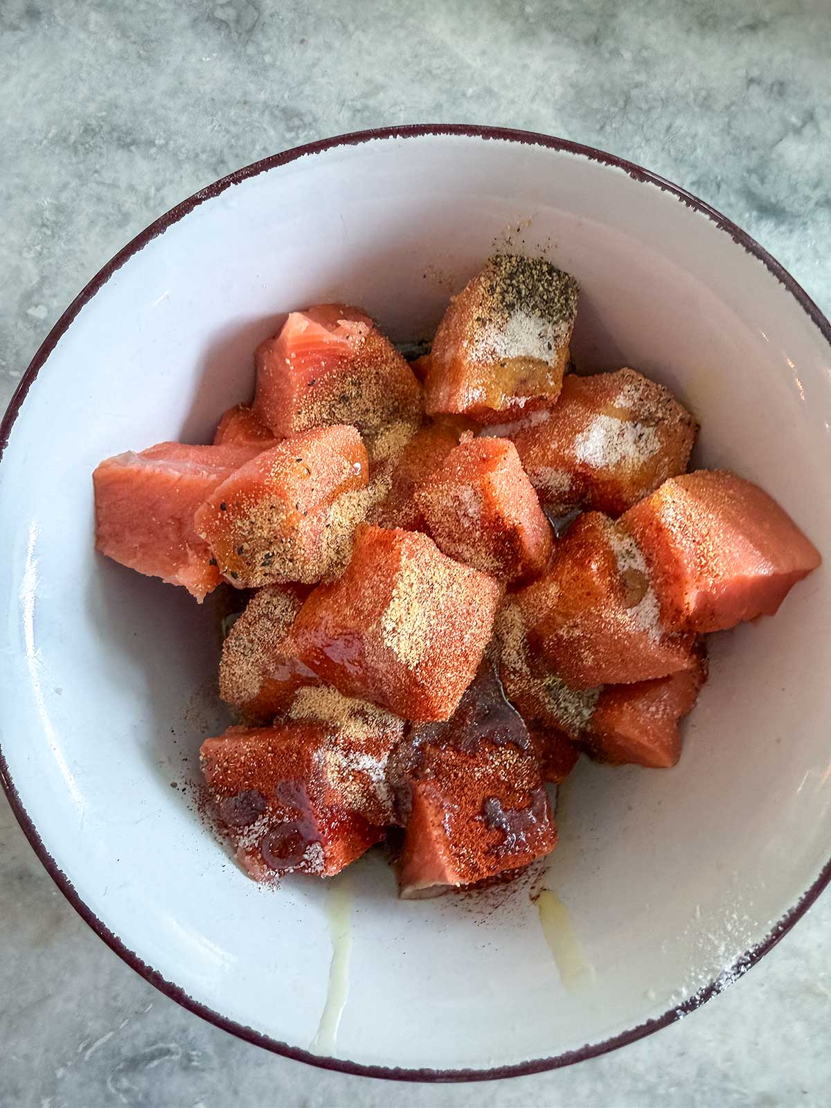 A white bowl filled with cubed salmon pieces sprinkled with salt, chili powder, and other spices, set on a gray marble surface—an inviting contrast to savory dishes like air fryer salmon bites.