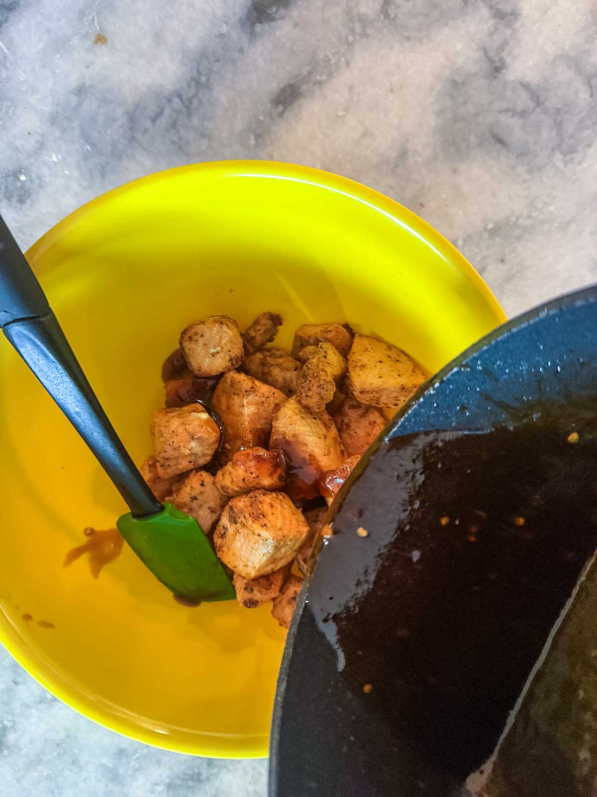 Cooked salmon pieces in a yellow bowl with a green spatula, while a dark sauce—similar to the glaze used for Honey Garlic Salmon—is being poured from a pan onto the chicken. Marble countertop in the background.