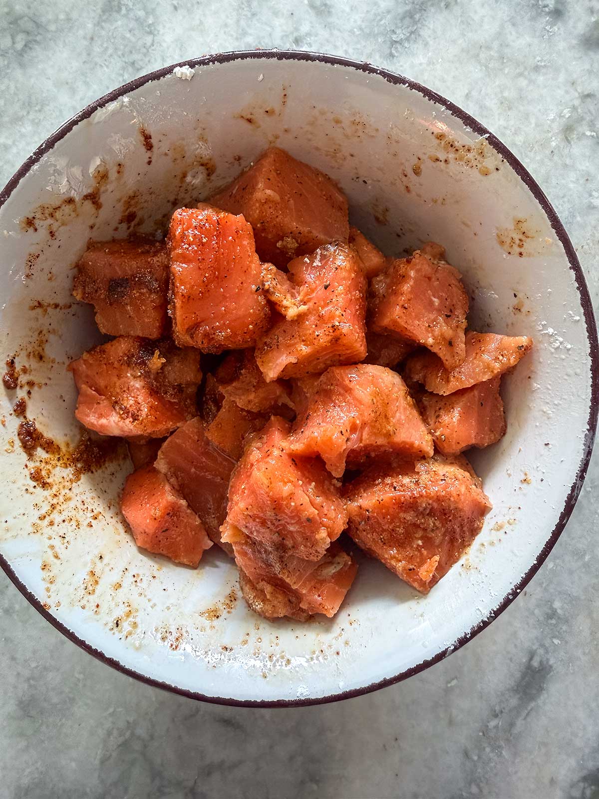 Chunks of raw salmon coated with seasonings and spices in a white bowl, placed on a gray marble surface—perfect for making delicious Honey Garlic Salmon Bites in the Air Fryer.